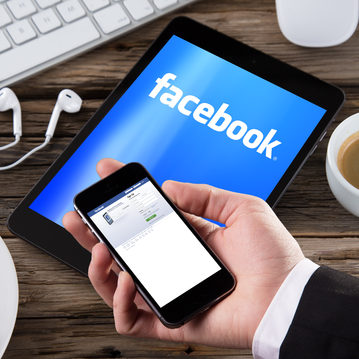 dreamstime_xs_38141139 hand holding phone over tablet screen with facebook open